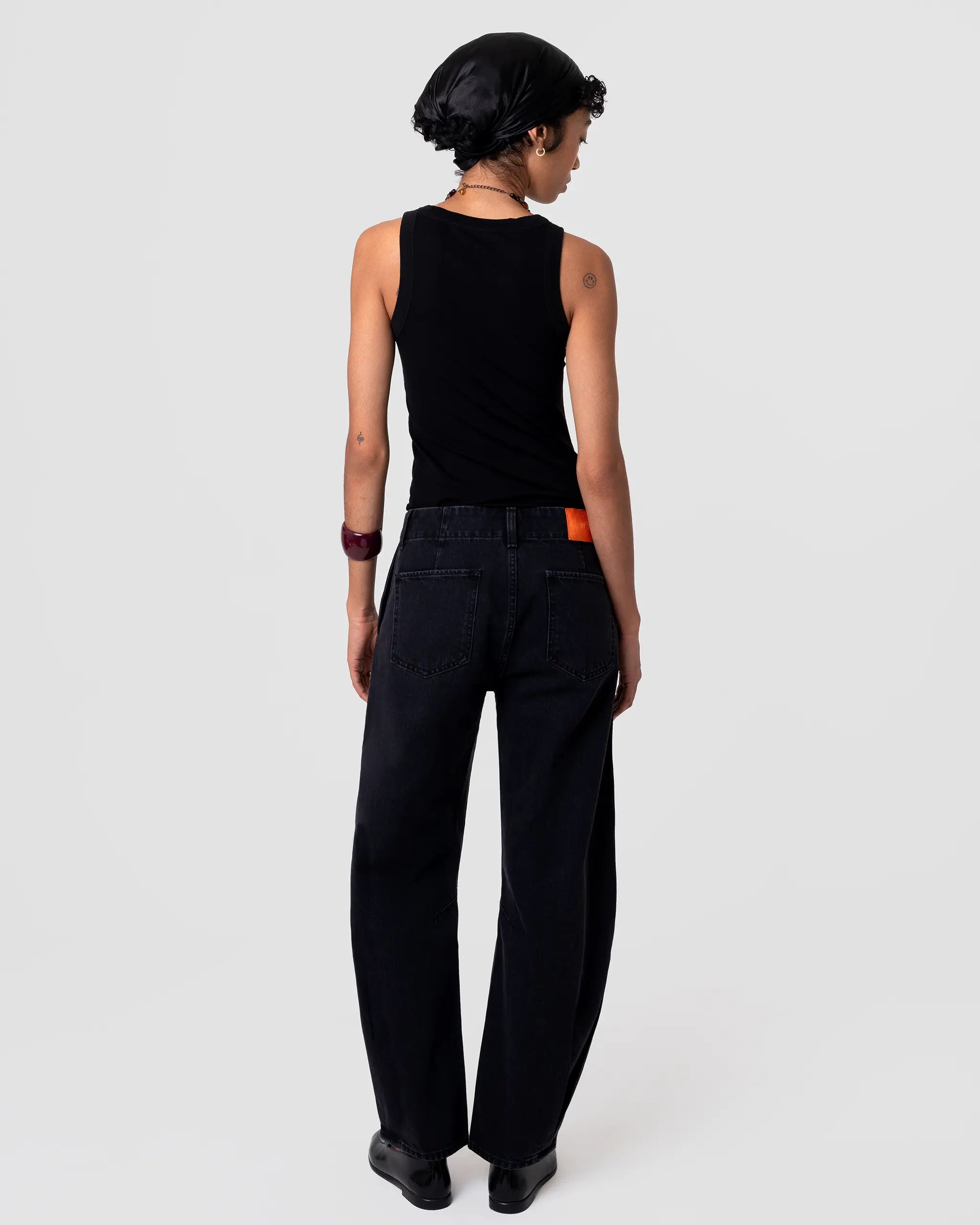 Rear view of Oketo Cut 103 Barrel Jean in deep black denim; relaxed drape, mid rise, barrel leg, orange back patch label visible. Pleated hem detail at cuff adds volume.