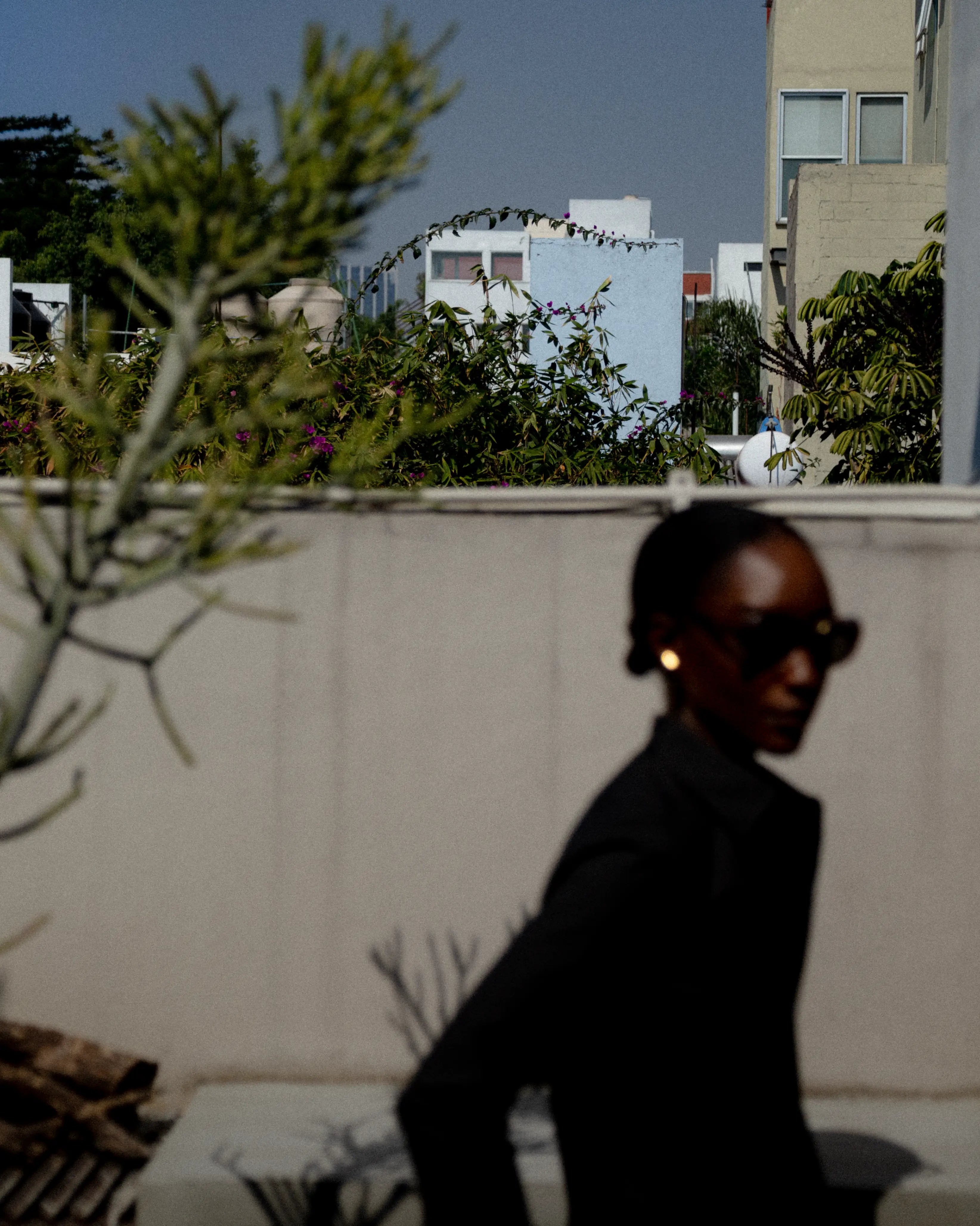 Foreground shows a blurred silhouette in dark denim; garment details obscured by focus on light urban backdrop and greenery.