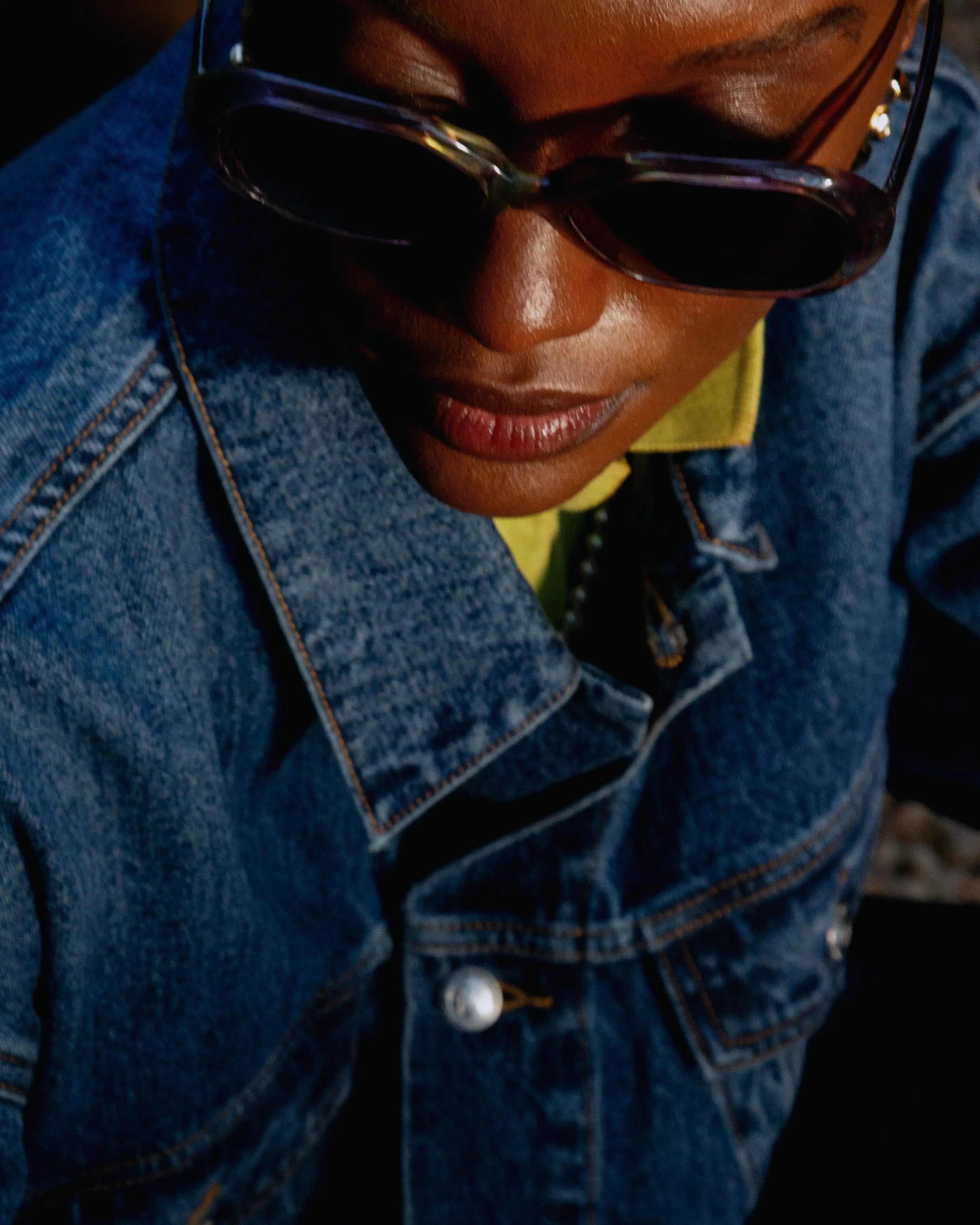 Close top view of a cropped indigo denim jacket; structured collar, soft fading, boxy fit over a yellow base layer.